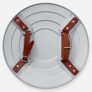 Puede incluir: Un escudo redondo de color plateado con una serie de surcos conc&eacute;ntricos. El escudo tiene correas de cuero marr&oacute;n y remaches met&aacute;licos para sujetarlo. La parte posterior del escudo es visible, mostrando los puntos de fijaci&oacute;n de las correas.