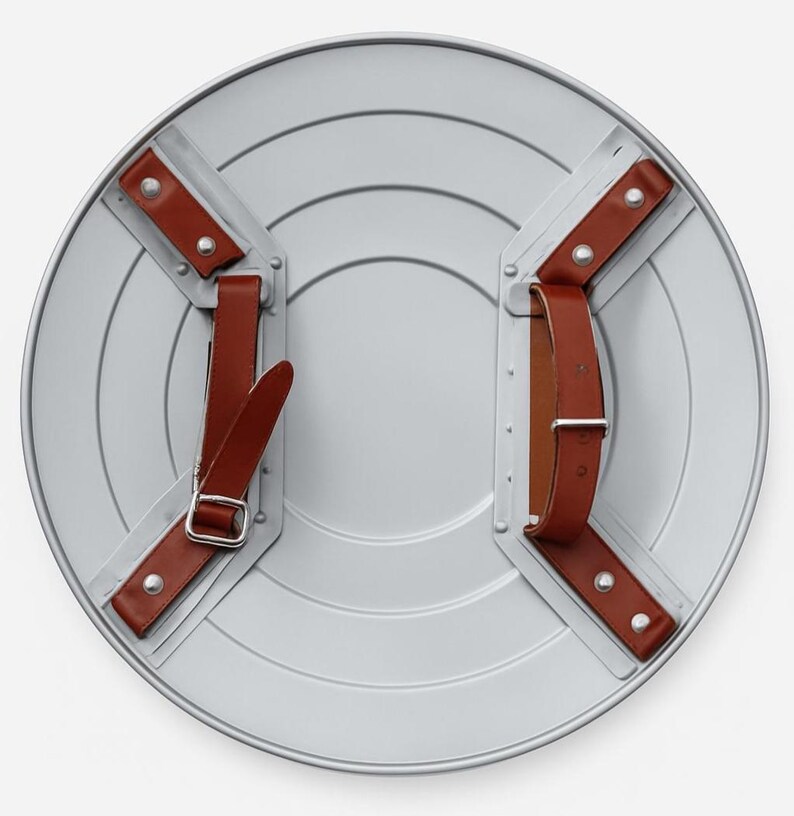 Puede incluir: Un escudo redondo de color plateado con una serie de surcos conc&eacute;ntricos. El escudo tiene correas de cuero marr&oacute;n y remaches met&aacute;licos para sujetarlo. La parte posterior del escudo es visible, mostrando los puntos de fijaci&oacute;n de las correas.
