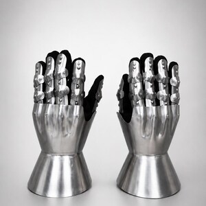 May include: A pair of articulated metal gauntlets featuring a polished silver finish. Each glove consists of a flared steel cuff, a hand plate, and segmented finger coverings with small rivets, all attached to a black fabric base for flexibility.