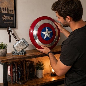 Puede incluir: Un escudo del Capit&aacute;n Am&eacute;rica con un dise&ntilde;o rojo, plateado y azul, con una estrella blanca en el centro. El escudo est&aacute; siendo sostenido por una persona. Tambi&eacute;n se ve el martillo de Thor y un p&oacute;ster de los Vengadores.