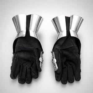 May include: A pair of black gloves featuring polished silver metal gauntlet cuffs and articulated metal plates along the outer edges. The gloves are constructed from a dark, textured fabric and include visible rivets securing the metal components.