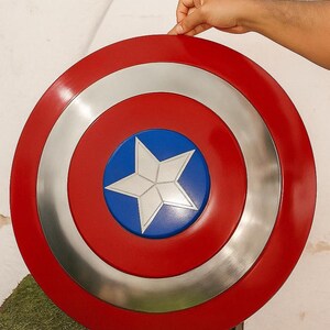 Puede incluir: Un escudo redondo con un borde exterior rojo, un anillo plateado y un centro azul. Una estrella blanca est&aacute; en el centro. El escudo est&aacute; sostenido por una mano.