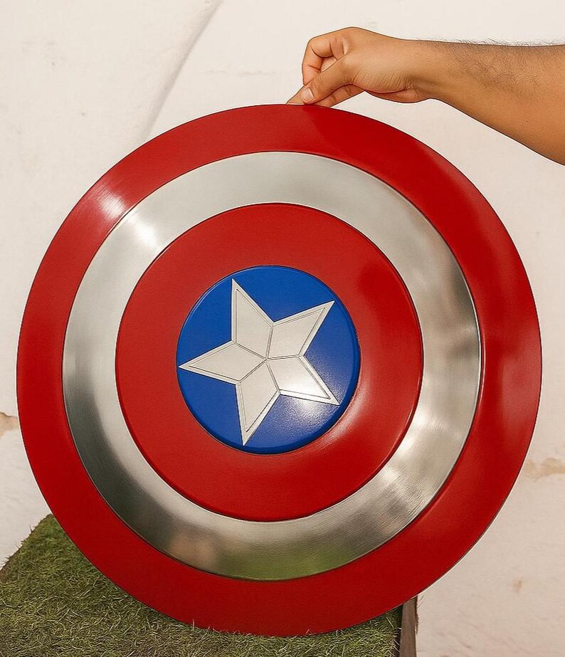 Puede incluir: Un escudo redondo con un borde exterior rojo, un anillo plateado y un centro azul. Una estrella blanca est&aacute; en el centro. El escudo est&aacute; sostenido por una mano.