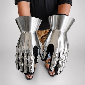 May include: A pair of articulated metal gauntlets featuring a polished silver finish. The gauntlets consist of a flared wrist cuff and segmented finger plates connected by small rivets, layered over a black fabric glove base for flexibility.