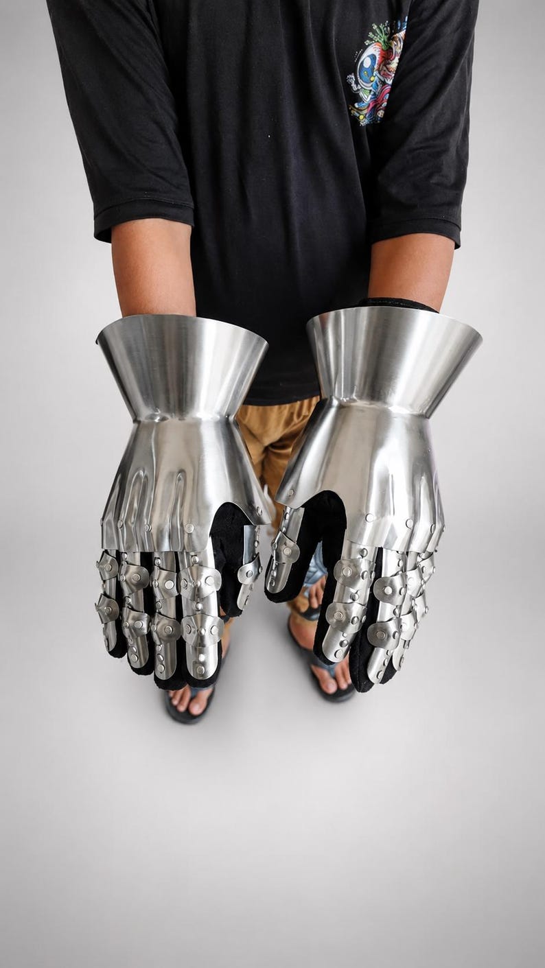 May include: A pair of articulated metal gauntlets featuring a polished silver finish. The gauntlets consist of a flared wrist cuff and segmented finger plates connected by small rivets, layered over a black fabric glove base for flexibility.