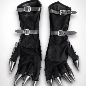 May include: A pair of black gloves featuring long, pointed metallic claws on each fingertip. The gloves are constructed from a soft, dark fabric and include two adjustable leather straps with silver-toned buckles across the forearm area.