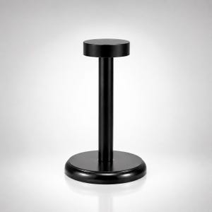 May include: A matte black display stand featuring a circular base, a vertical cylindrical post, and a flat circular top platform. The structure is uniform in color and has a smooth finish, designed to hold items securely in an upright position.