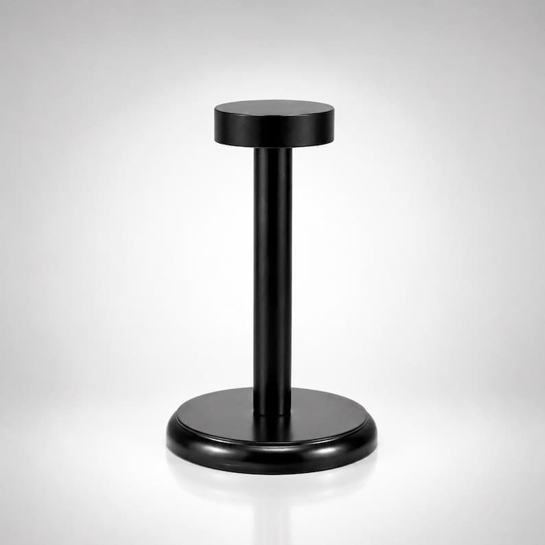 May include: A matte black display stand featuring a circular base, a vertical cylindrical post, and a flat circular top platform. The structure is uniform in color and has a smooth finish, designed to hold items securely in an upright position.