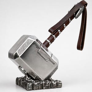 Puede incluir: Un martillo plateado con intrincados grabados en la cabeza, apoyado sobre una base texturizada de color gris oscuro. El mango est&aacute; envuelto en cuero marr&oacute;n con detalles plateados y una correa a juego.