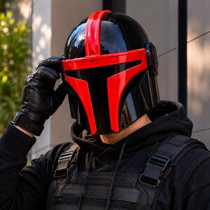 May include: A glossy black helmet featuring a prominent red T-shaped visor and a central red stripe running over the top. The helmet has a smooth, reflective finish and is worn with a black hooded sweatshirt and a black tactical vest.