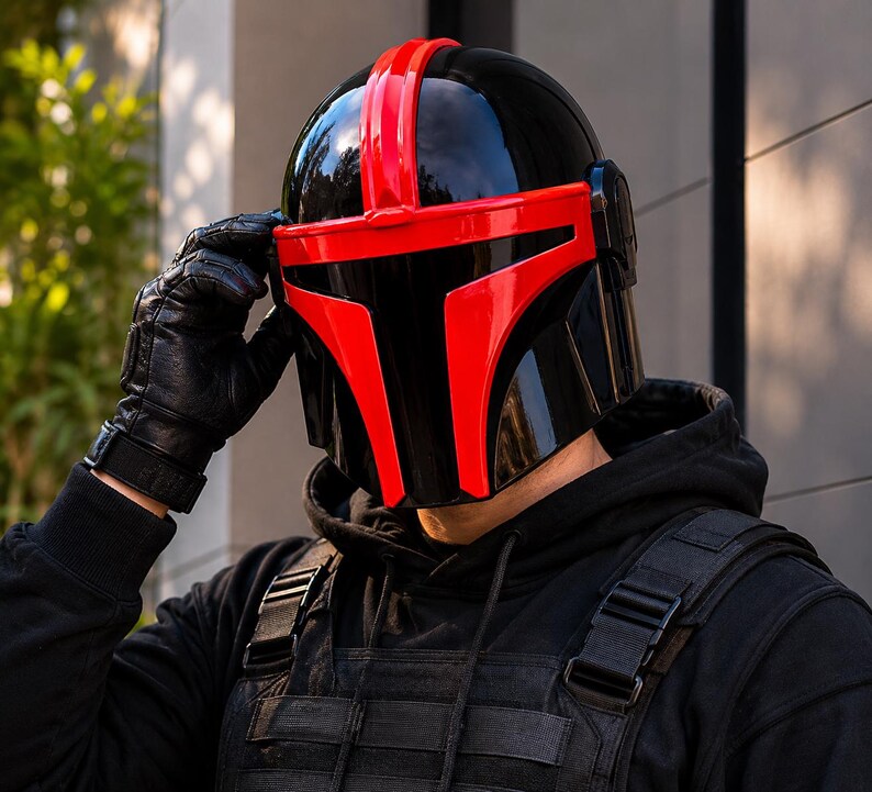 May include: A glossy black helmet featuring a prominent red T-shaped visor and a central red stripe running over the top. The helmet has a smooth, reflective finish and is worn with a black hooded sweatshirt and a black tactical vest.