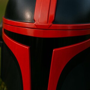 May include: A full-face helmet featuring a matte black finish with glossy red accents. The design includes a prominent red T-shaped visor frame and a vertical red stripe running along the center of the dome, creating a bold contrast.