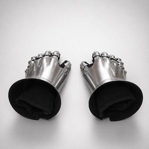 May include: A pair of articulated metal gauntlets featuring polished steel plates covering the fingers and back of the hand, attached to black fabric cuffs. The armor pieces are connected with rivets and designed to protect the hands and wrists.