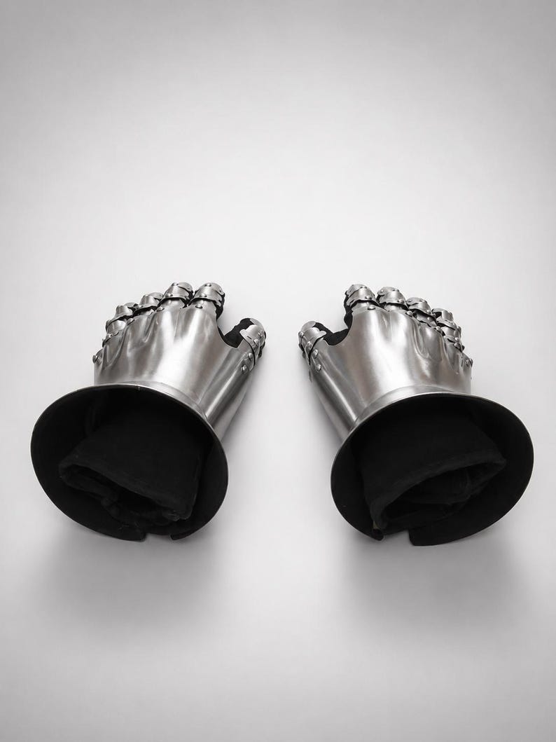 May include: A pair of articulated metal gauntlets featuring polished steel plates covering the fingers and back of the hand, attached to black fabric cuffs. The armor pieces are connected with rivets and designed to protect the hands and wrists.