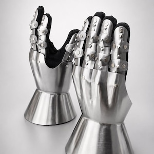 May include: A pair of articulated metal gauntlets featuring polished steel plates over a black fabric base. Each gauntlet consists of a flared cuff, a solid hand plate, and segmented finger coverings secured with small rivets for flexible movement.