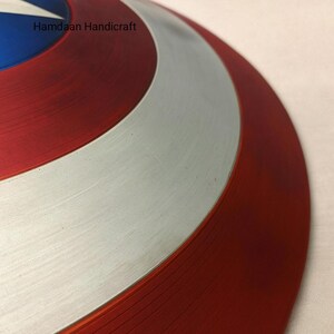 May include: A close-up of a red, white, and blue shield with a star in the center. The shield has a circular design with a silver band and the text "Hamdaan Handicraft". The shield is a replica of a superhero's shield.