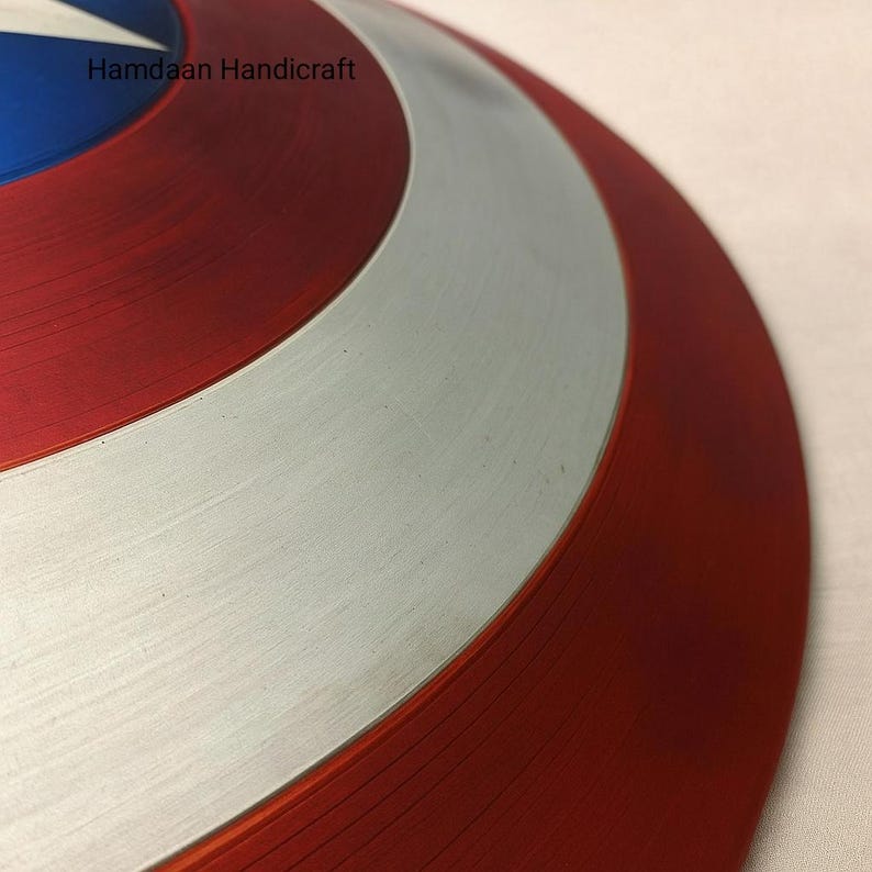 May include: A close-up of a red, white, and blue shield with a star in the center. The shield has a circular design with a silver band and the text "Hamdaan Handicraft". The shield is a replica of a superhero's shield.
