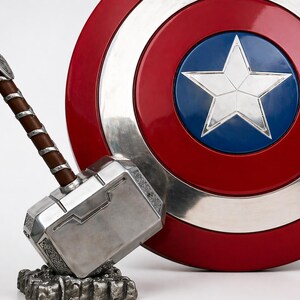 Puede incluir: Un martillo de Thor plateado y marr&oacute;n descansa sobre una base rocosa junto a un escudo del Capit&aacute;n Am&eacute;rica rojo, blanco y azul. El escudo tiene un borde plateado y una estrella blanca sobre un c&iacute;rculo azul en el centro. El martillo tiene un mango marr&oacute;n.