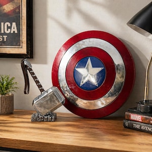 Puede incluir: Un escudo del Capit&aacute;n Am&eacute;rica con una estrella plateada sobre un c&iacute;rculo azul, rodeado por un anillo plateado y un anillo exterior rojo. Junto a &eacute;l, el martillo de Thor, Mjolnir, con mango marr&oacute;n y cabeza plateada. Sobre una superficie de madera.
