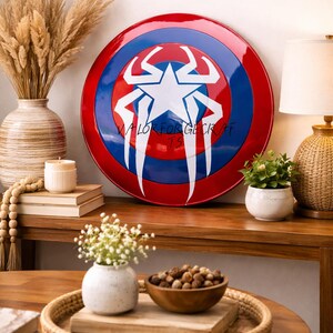 May include: A round shield with a red border, blue center, and a white starburst design. The shield has the text "VALOR FOR GECK AF TS" in white. The shield is displayed on a wooden surface with decorative items.