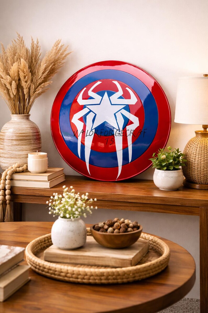 May include: A round shield with a red border, blue center, and a white starburst design. The shield has the text "VALOR FOR GECK AF TS" in white. The shield is displayed on a wooden surface with decorative items.