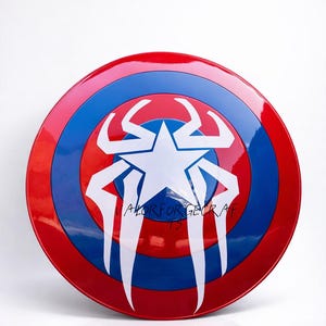 May include: A red, white, and blue shield with a star and spider-like design. The shield has a glossy finish and the text "VALOR FOR GECRAF 13" is written on it. The shield is a prop or collectible.