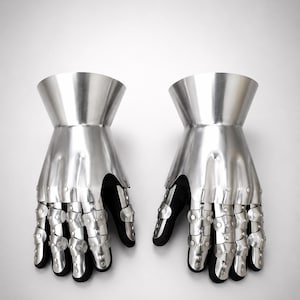 May include: A pair of articulated metal gauntlets featuring a polished silver finish. Each gauntlet consists of a flared cuff, a solid hand plate, and segmented finger coverings connected by rivets, all mounted over a black fabric base.
