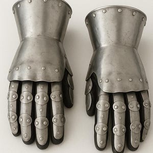 May include: A pair of articulated metal gauntlets featuring a polished steel finish. Each glove consists of a wide cuff with rivets, a hand plate, and segmented finger coverings with small oval metal plates secured by rivets over a dark base.