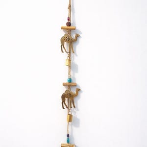 May include: A decorative hanging ornament with three brass-coloured camel silhouettes, small bells, and wooden accents. The ornament is suspended by a natural twine cord, with colourful beads adding visual interest. The design has an ethnic or desert theme.