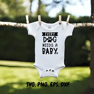 Op de afbeelding: Witte babybody met de zwarte tekst "EVERY DOG NEEDS A BABY". De body hangt aan een waslijn met houten knijpers tegen een vage groene achtergrond.