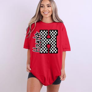 May include: A red t-shirt featuring the word "Hawks" in a script font and a large, checkered "H" design. The shirt is a short-sleeved, casual style.