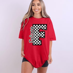 Puede incluir: Una camiseta roja con un gran diseño de "F" a cuadros y la palabra "Falcons" en una fuente cursiva. La camiseta es de manga corta y tiene un corte holgado. El diseño es en blanco y negro.