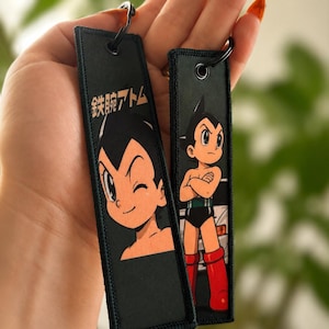 May include: Two rectangular black fabric keychains featuring Astro Boy graphics. One keychain shows a close-up of Astro Boy winking with the Japanese text 鉄腕アトム, while the other displays a full-body image of the character with arms crossed.