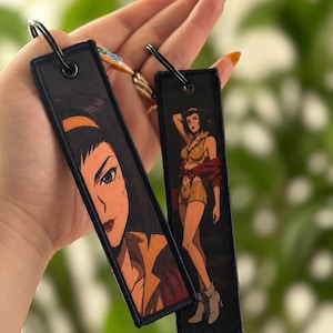 May include: Two rectangular black fabric keychains featuring anime-style illustrations of a character with dark hair. One keychain shows a close-up of the character's face, while the other displays a full-body view of the character in a yellow outfit.