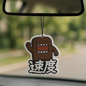 May include: A brown Domo-kun air freshener with a white border hangs from a car's rearview mirror. The air freshener features Domo-kun's face with an open mouth and the Japanese characters for "speed" in white.