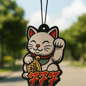 May include: A car air freshener featuring a cartoon Maneki-neko cat with a raised paw, holding a gold coin with Japanese characters. The design includes the number 777 in red, with a black background and a black string for hanging.