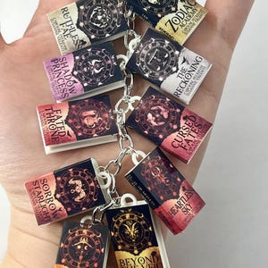 Può includere: Un portachiavi con ciondoli a forma di libri in miniatura. Ogni ciondolo presenta un design di copertina unico con titoli come "Ruthless Fae", "Zodiac Academy" e "Shadow Princess". I libri sono collegati da una catena d'argento, creando un accessorio stravagante.