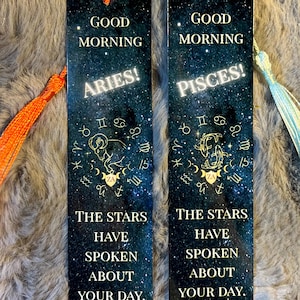 May include: Two rectangular bookmarks with tassels. One bookmark says "Good Morning Aries!" and the other says "Good Morning Pisces!". Both have a starry night sky background with zodiac symbols and the text "The stars have spoken about your day."