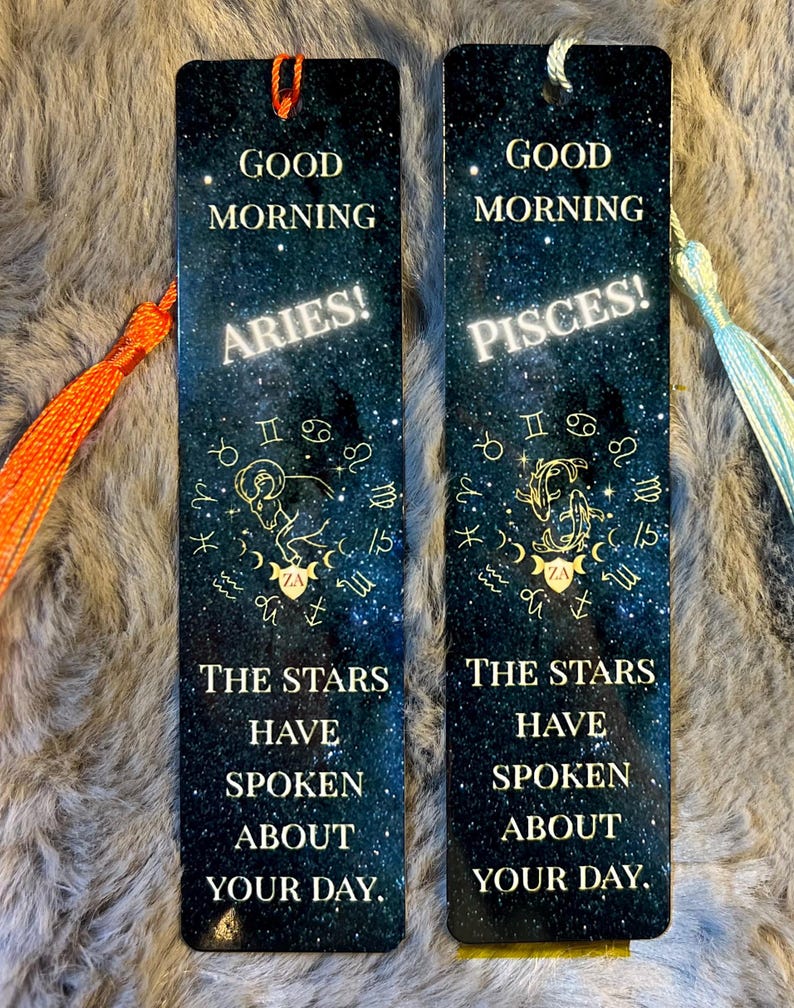 May include: Two rectangular bookmarks with tassels. One bookmark says "Good Morning Aries!" and the other says "Good Morning Pisces!". Both have a starry night sky background with zodiac symbols and the text "The stars have spoken about your day."