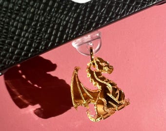 May include: A gold-colored dragon charm with detailed wings and body, hanging from a clear plastic clasp. The charm is set against a pink background, with a shadow cast to the left. A black textured surface and a red and white sticker are visible at the top.