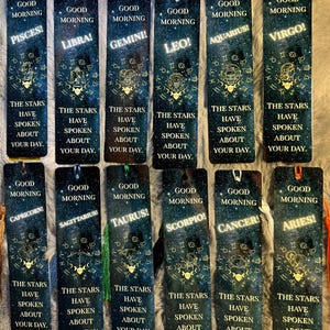 May include: A collection of twelve rectangular bookmarks, each featuring a different zodiac sign. The bookmarks have a dark blue background with a starry design and gold accents. Each bookmark has a tassel and the text "Good Morning" and a zodiac sign.