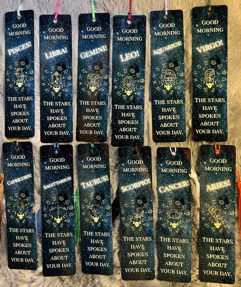 May include: A collection of twelve rectangular bookmarks, each featuring a different zodiac sign. The bookmarks have a dark blue background with a starry design and gold accents. Each bookmark has a tassel and the text "Good Morning" and a zodiac sign.