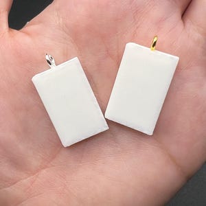 May include: Two rectangular white pendants with smooth surfaces. One pendant features a silver-toned metal bail at the top, while the other pendant features a gold-toned metal bail at the top. Both items are simple, flat, and rectangular in shape.