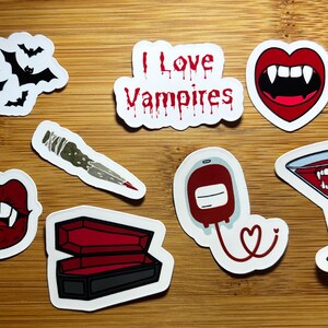 May include: A collection of vampire-themed stickers on a wooden surface. Designs include bats, the text "I Love Vampires", vampire fangs, a stake, a coffin, a blood bag, and a martini glass with fangs.