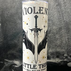 Op de afbeelding: Een witte beker met de woorden "VIOLENT LITTLE THINGS" en "I WILL NOT DIE TODAY" in zwarte letters. Het ontwerp bevat een zwart zwaard met vleermuisvleugels en gouden sterren.