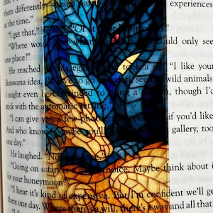 May include: A bookmark featuring a stained-glass style dragon design. The dragon is depicted in shades of blue, with a yellow and orange underbelly. The bookmark is set against a backdrop of text from a book.
