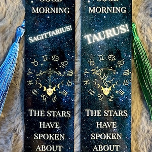 May include: Two rectangular bookmarks with a starry night sky design. One features "Sagittarius!" and the other "Taurus!" in white text. Both have gold zodiac symbols and the text "Good Morning" and "The Stars Have Spoken About Your Day."