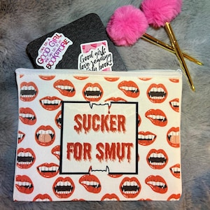 May include: A white zippered pouch with a repeating pattern of red vampire fangs and lips. The pouch has the text "SUCKER FOR SMUT" in a black outlined box. A black notebook with stickers and two pink pom-pom pens are also in the image.