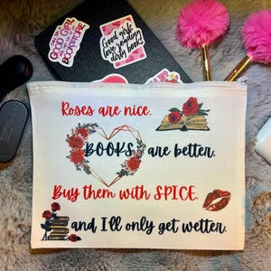 May include: A white canvas pouch with the text "Roses are nice. BOOKS are better. Buy them with SPICE. and I'll only get wetter." is surrounded by floral and book graphics. The pouch is next to a black notebook with stickers, pens, and other accessories.