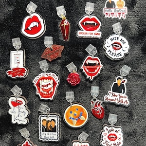 May include: A selection of acrylic earrings with diverse designs, such as vampire fangs, coffins, roses, and phrases like "Bite Me Please" and "I Love Vampires." The earrings feature red, white, and black colours.